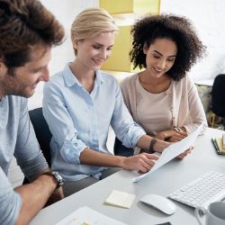 Drei Personen sitzen mit Zetteln vor PC.
