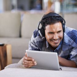 Mann liegt mit Kopfhoerern und Tablet auf dem Sofa und lacht.