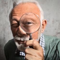 Ein alter Mann hält eine Lupe vor sein Auge in die Kamera.