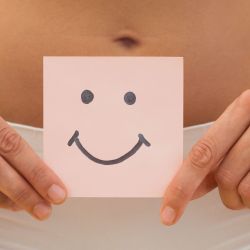 Eine junge Frau hält ein Stück Papier mit einem Smiley drauf über ihren Bauch.
