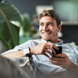 Mann sitzt auf der Couch und hält lächelnd ein Smartphone in der Hand.
