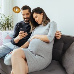Eine schwangere Frau und ihr Mann sitzen auf dem Sofa und schauen zusammen auf ein Smartphone.