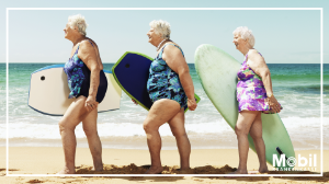 Ältere Damen laufen mit Surfbrettern unter dem Arm am Strand entlang.