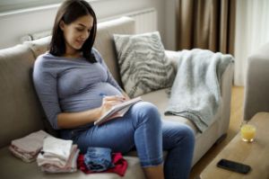 Eine Schwangere sitzt auf der Couch und prüft Ihre Checkliste zur Geburtsvorbereitung.