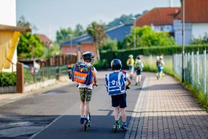 Zwei Schulkinder mit Schulranzen und Helmen fahren mit ihren Rollern auf der Strasse, man sieht die beiden von hinten.