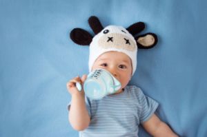 Kleinkind mit Kuh-Mütze trinkt im Liegen selbst aus einer Flasche.