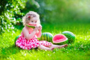 Kleines Maedchen sitzt auf einer Wiese mit Melonen und isst ein kleines Stück Melone.