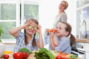Zwei Kinder machen mit Gemüse Quatsch und haben Spaß in der Küche. Die Mutter lacht im Hintergrund.