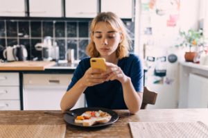 Teenager-Maedchen sitzt am Esstisch in der Kueche, sie schaut auf das Smartphone, vor ihr steht ein Teller mit Essen.