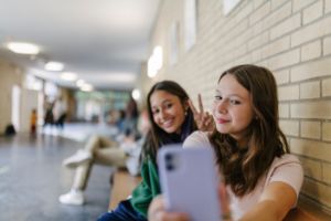 Zwei Teenager-Mädchen sitzen im Flur in der Schule auf einer Bank und machen eine Selfie mit dem Smartphone.