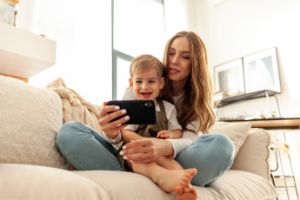 Mutter hat ihr Kleinkind auf dem Schoss, beide schauen auf das Smartphone, das die Mutter in der Hand hält, und lachen.