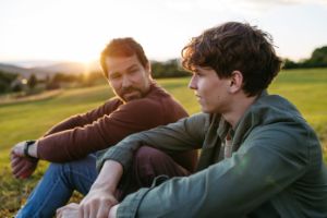Vater und Teenager-Sohn sitzen draußen nebeneinander, der Sohn sieht nachdenklich aus, der Vater schaut ihn an.