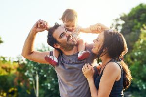 Vater trägt Kleinkind auf Schultern, Mutter schaut beide an, alle lachen und sind fröhlich.