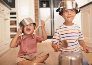 Zwei Kinder sitzen mit Töpfen auf dem Kopf und einem Kochlöffel in der Hand auf dem Küchenboden vor einem umgedrehten Kochtopf.