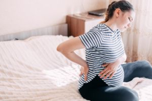 Schwangere sitzt auf Bett, hält sich Bauch und Rücken, hat Beschwerden.