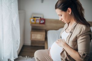 Schwangere Frau sitzt auf Bettkante, hält ihren Bauch und blickt sorgenvoll.