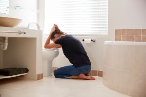 Junge Frau kniet über der offenen Toilette, hält sich den Kopf, muss sich übergeben.