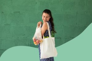 Eine junge Frau schaut lächelnd auf Ihre zwei Einkaufstüten voller Obst und Gemüse über der Schulter.