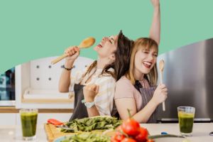Zwei junge Frauen stehen mit Kochlöffeln als Mikrofone singend in der Küche. Auf dem Tisch stehen zwei Smoothies und viel Gemüse.