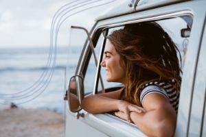 Eine junge Frau sitzt in einem kleinen Campingbus am Stand und schaut lächelnd auf das Meer.