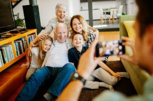 Ein Rentnerpärchen posiert im Wohnzimmer gut gelaunt mit seinen 3 Enkeln für ein Foto. Ein Mann nimmt das Foto mit einem Smartphone auf.