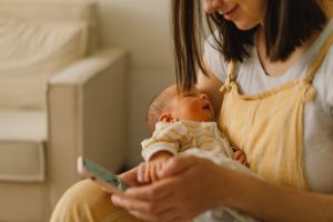 Mutter mit Säugling auf dem Arm schaut sich etwas auf ihrem Smartphone an.