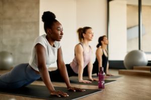 Drei Frauen führen eine Pilates-Übung auf einer Sport-Matte in einem Kursraum aus.