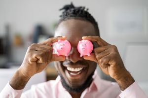 Mann mit breitem Grinsen hält sich jeweils ein kleines rosa Sparschwein vor seine Augen.