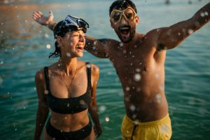 Frau steht neben einem Mann, der mit seinen Händen Wasser hinauf spritzt, im Meer.