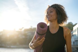 Frau steht in der Natur mit der Morgensonne im Rücken und trägt eine Yogamatte unter ihrem Arm
