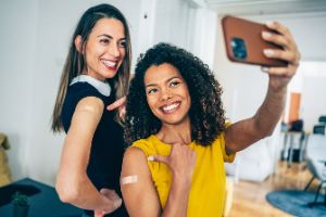 Zwei junge Frauen machen ein Selfi und zeigen auf ihren geimpften Arm mit Pflaster.