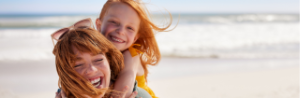 Mutter trägt Kind auf dem Rücken am Strand, beide lachen.