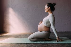 Eine schwangere Frau sitzt auf den Knien auf einer Gymnastikmatte und hält die Hand auf ihren Bauch.