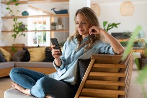 Junge Frau sitzt auf dem Sofa und schaut lächelnd auf ihr Smartphone.