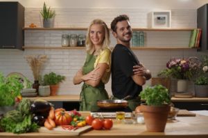 Frau und Mann stehen Rücken an Rücken in Küche, lachen in die Kamera. Auf der Arbeitsfläche liegt viel Gemüse.