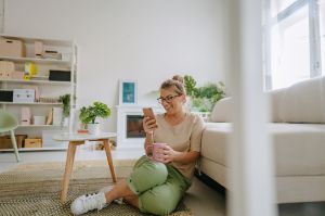 Eine Frau sitz in ihrem Wohnzimmer mit einer Tasse in der Hand auf dem Fußboden und schaut lachend auf ihr Smartphone.