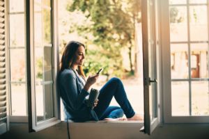 Eine junge Frau sitzt mit einem Thermobecher in der Hand im Fensterrahmen und schaut lächelnd auf ihr Smartphone.