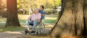 Frau schiebt älteren Mann im Rollstuhl durch einen Park.