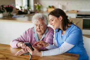 Eine junge Frau sitzt mit einer älteren Frau am Tisch und erklärt ihr etwas am Smartphone.