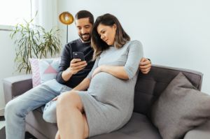 Eine schwangere Frau und ihr Mann sitzen auf dem Sofa und schauen zusammen auf ein Smartphone.