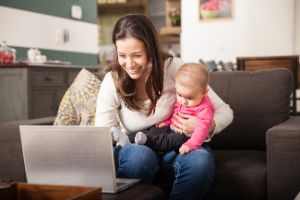 Frau mit Baby auf dem Schoß schaut lächelnd auf den Bildschirm eines Laptops.