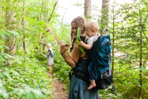 Eine Frau geht mit ihren Kindern im Wald spazieren Das Kleinkind in einer Trage auf dem Rücken freut sich über ein großes Blatt.