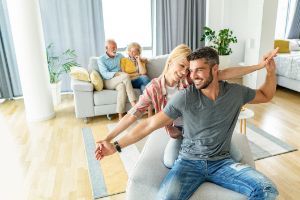 Eine Frau umarmt lachend ihren Freund, der an Tisch vor einem Notebook sitzt.