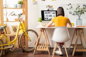 Eine Frau sitzt zu Hause am Schreibtisch und schaut auf ihren Computerbildschirm. Neben ihr steht ein gelbes Fahrrad.