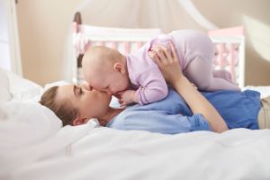 Eine Frau liegt auf einem Bett, hält ihr Baby auf dem Bauch und gibt ihm einen Kuss auf die Stirn.