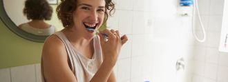 Eine junge Frau mit Locken steht im Bad und putzt sich fröhlich die Zähne. 