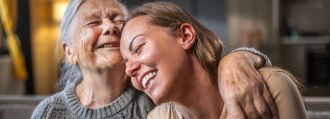 Eine ältere und eine jüngere Frau sitzen auf dem Sofa, umarmen sich und lachen dabei.