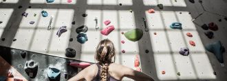 Eine Frau steht vor einer Boulderwand und schaut hoch.