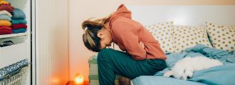 Eine junge Frau sitzt gekrümmt vor Bauchschmerzen auf dem Bett.