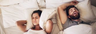 Ein junges Paar liegt im Bett, der Mann schnarcht und die Frau hält sich die Ohren zu.
